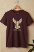 A maroon short-sleeve t-shirt hanging on a wooden hanger against a textured beige wall. The shirt features a cream-colored graphic of an angel with outstretched wings and the words 'DIVINE GRACE' printed below.