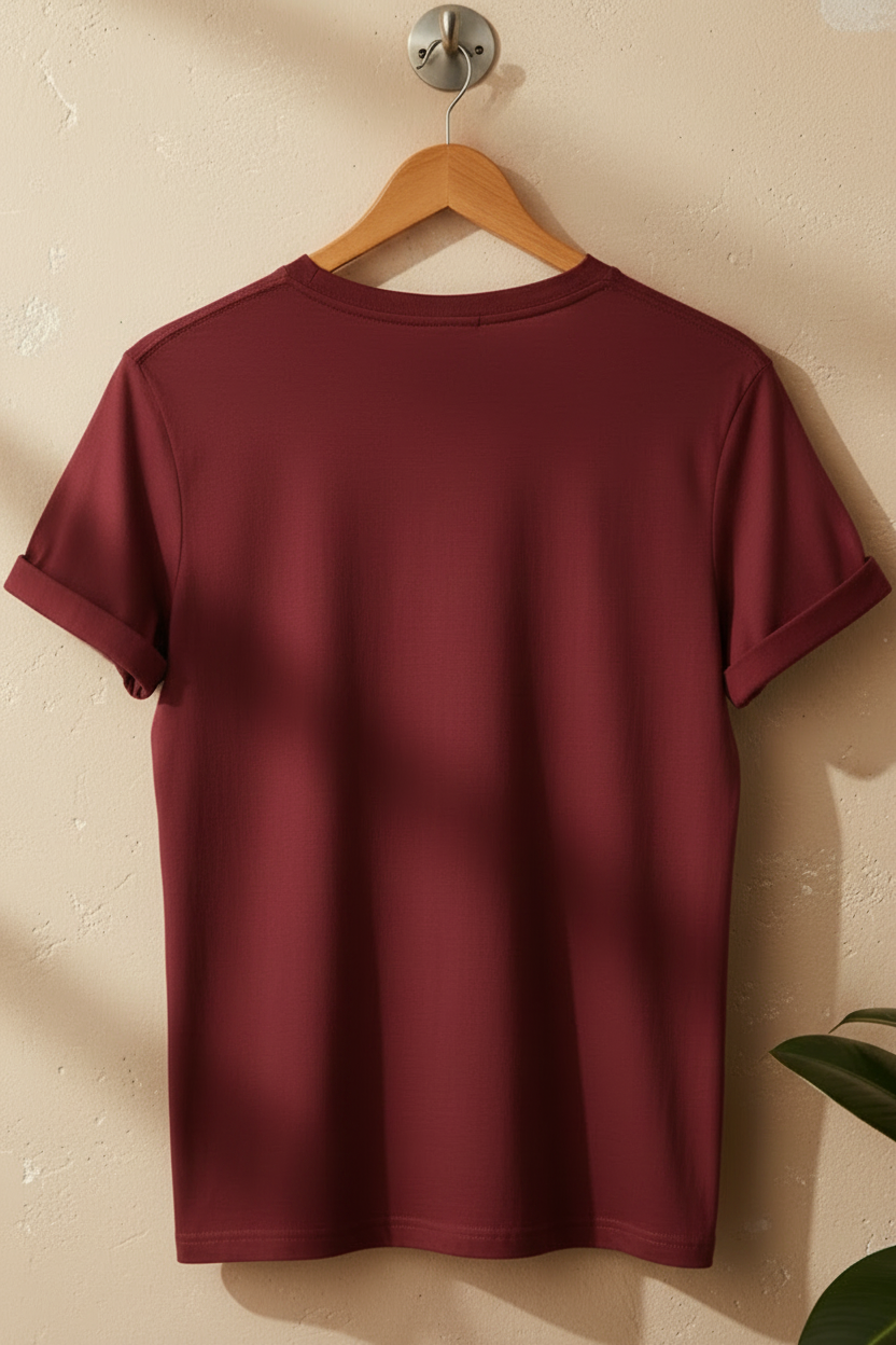 A short-sleeve crewneck t-shirt in a deep burgundy red, hanging on a wooden hanger against a textured beige wall with soft diagonal shadows.