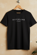 A black short-sleeve crewneck t-shirt hanging on a wooden hanger against a textured beige wall. The shirt features the word 'DISCIPLINE' printed in white across the chest, with smaller text underneath reading 'DO IT NOW.