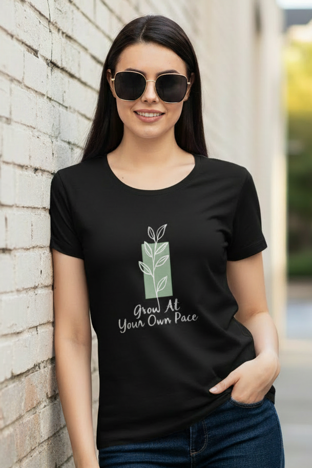 A woman wearing a black short-sleeve t-shirt with a green gift box graphic and the text 'Good At Your Own Pace' stands against a light brick wall.