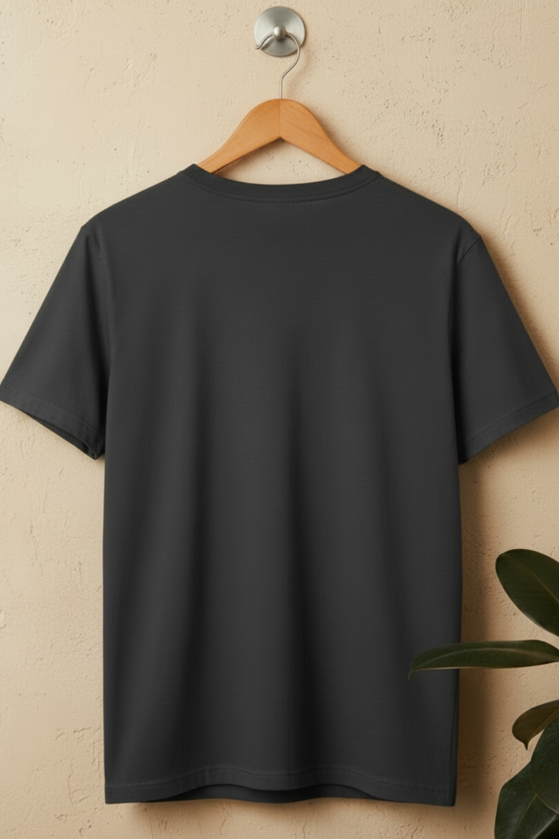 A plain dark charcoal gray short-sleeve t-shirt hanging on a wooden hanger against a textured beige wall, with a green plant leaf visible in the lower right corner.